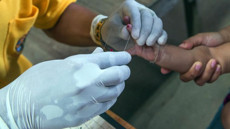 A healthcare worker with gloved hands draws blood from a child's finger.